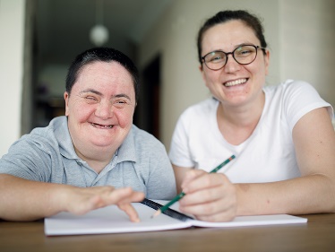 A person is helping someone else with writing in a booklet. Both are smiling. 